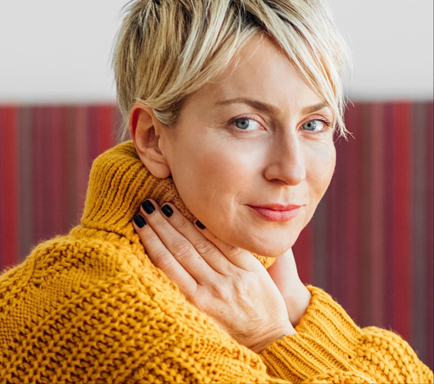 A woman in a yellow sweater, hand at her collar, looking at the camera